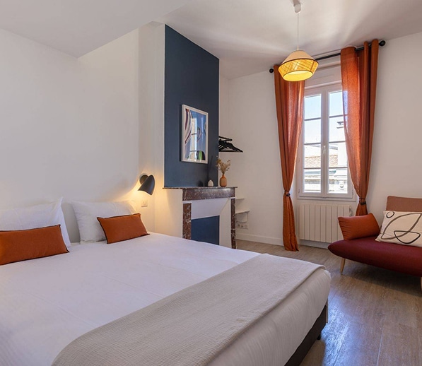 Chambre d'hôtel moderne avec lit double blanc, mur d'accent bleu nuit et cheminée décorative. Coussins et rideaux terracotta.