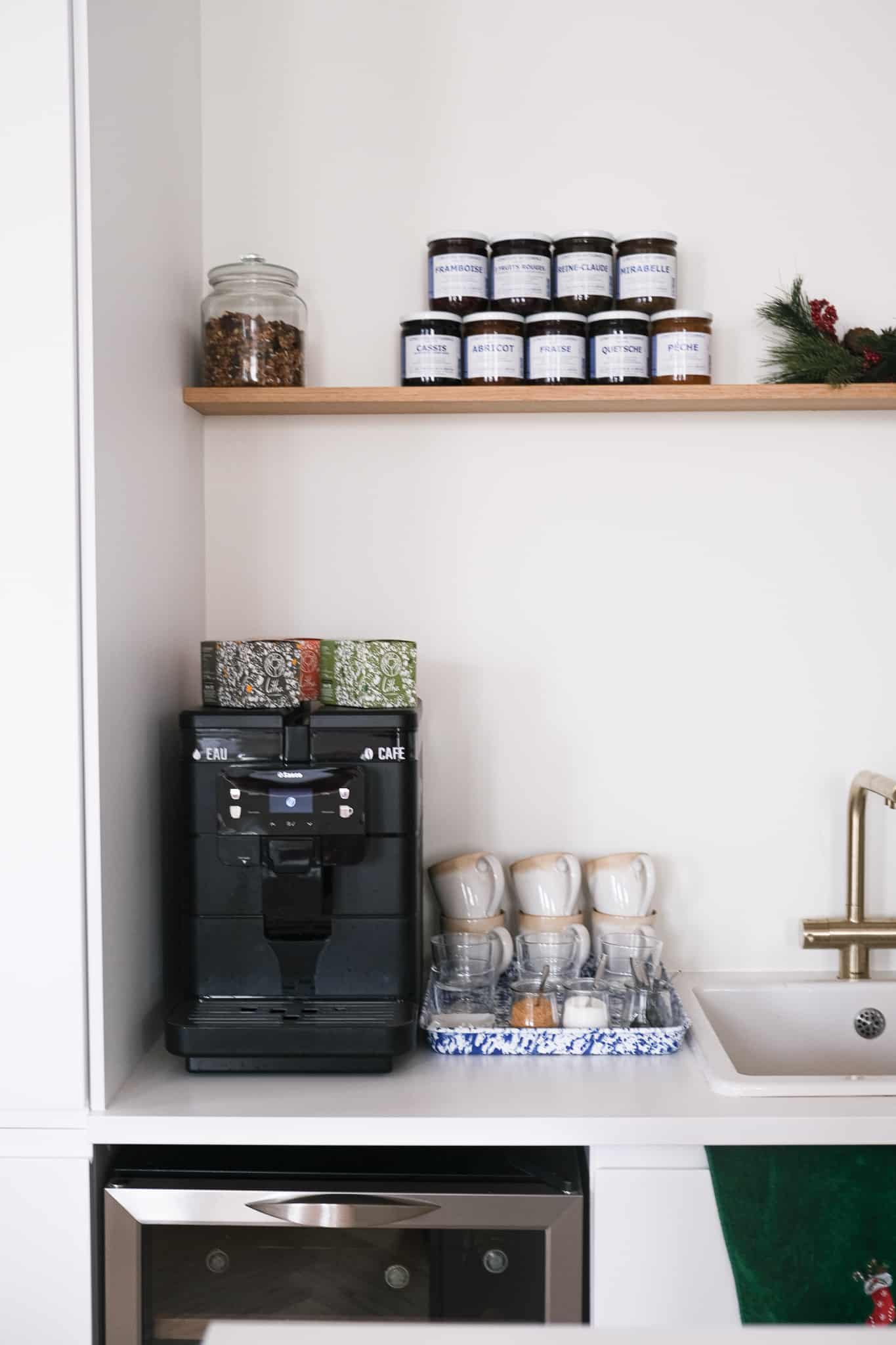 Cuisine moderne organisée. Station café noire, mugs empilés, évier laiton. Étagère en bois avec 11 pots de confiture (Fraise, Cassis, etc.) et granola.