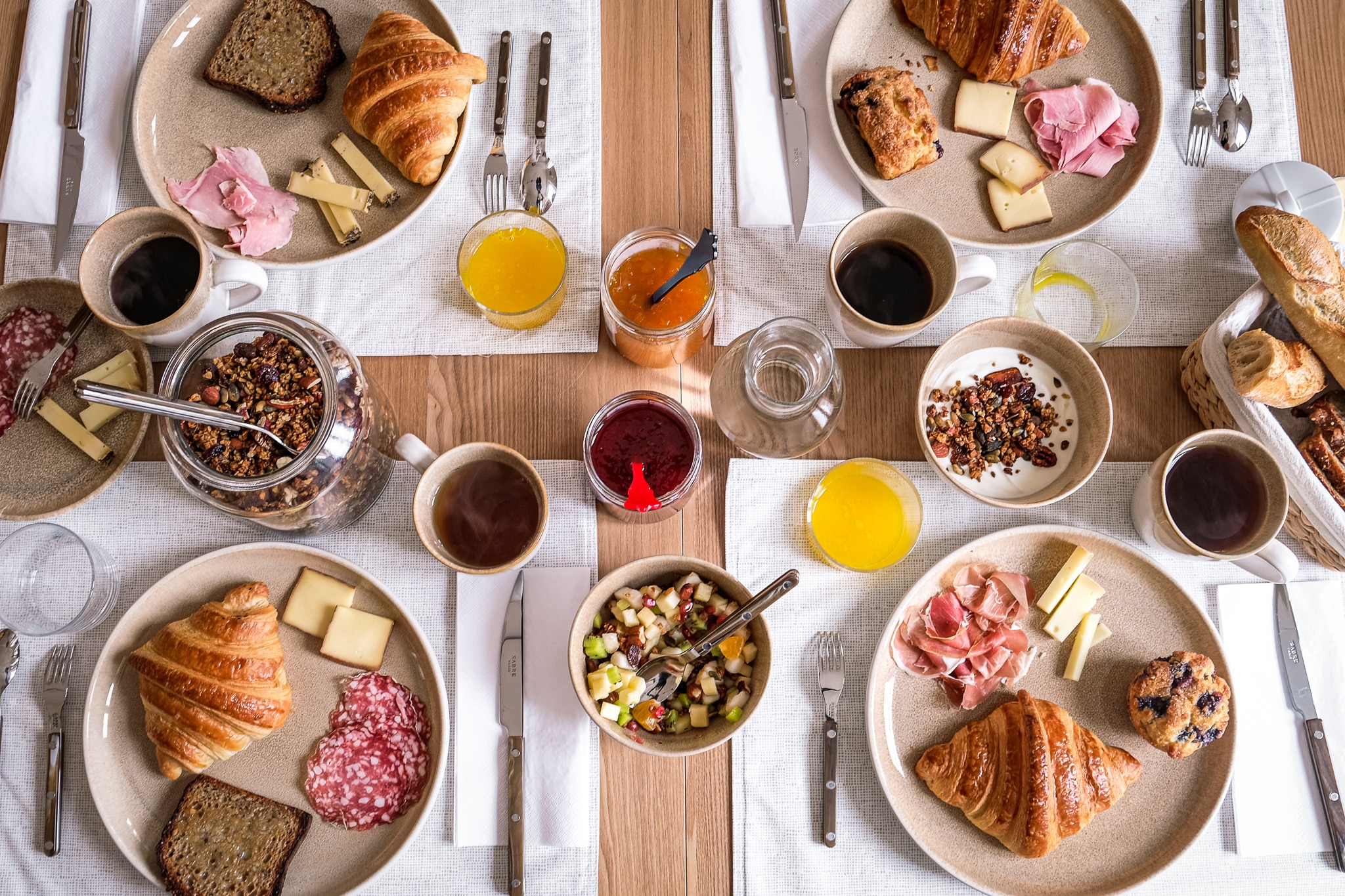 Table de brunch luxueuse vue du dessus : croissants, jambon, salami, fromages, café, jus d'orange, granola et confitures.